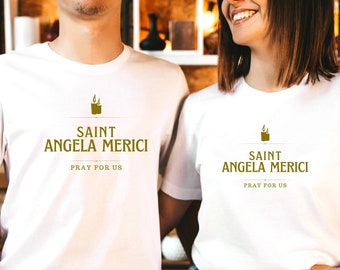 Könnte beinhalten: Zwei weiße T-Shirts mit einem goldenen Grafikdesign. Die Grafik zeigt eine Kerze mit Flammen und den Text "Saint Angela Merici Bete für uns".