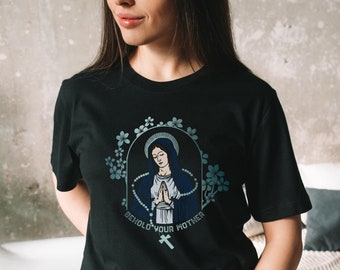 以下が含まれることがあります： 青いローブを着た女性と、ロザリオと花が描かれたグラフィックがプリントされた黒いTシャツ。画像の下には「Behold your mother」というテキストがプリントされています。