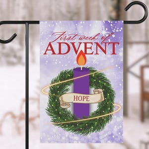 May include: A small garden flag featuring a purple Advent candle with a flame, surrounded by a green wreath and the word "HOPE" on a banner. The flag reads "First week of ADVENT" in red script against a lavender background.