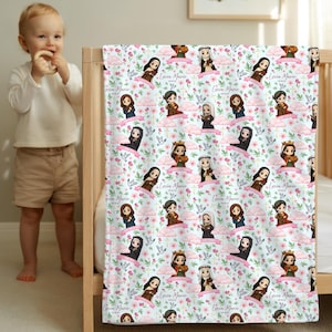May include: A white baby blanket with a repeating pattern of cartoon figures, pink flowers, and the text "Livia Marie" on a pink banner. The blanket is draped over a wooden crib.
