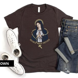 May include: A brown t-shirt with a graphic of a woman in a blue robe with a gold rosary and the text "Behold Your Mother".