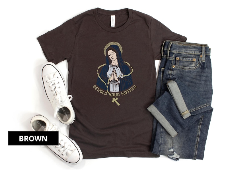 May include: A brown t-shirt with a graphic of a woman in a blue robe with a gold rosary and the text "Behold Your Mother".