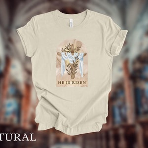 May include: A beige t-shirt with a watercolor illustration of a wooden cross with a white cloth draped over it. The cross is surrounded by green leaves and the text "HE IS RISEN" is printed below the cross.