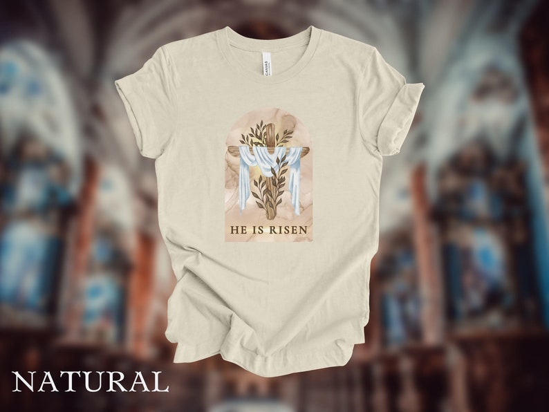 May include: A beige t-shirt with a watercolor illustration of a wooden cross with a white cloth draped over it. The cross is surrounded by green leaves and the text "HE IS RISEN" is printed below the cross.