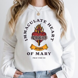 May include: White sweatshirt with a graphic print of a flaming heart with a sword and roses. The text around the heart reads "Immaculate Heart of Mary" and "Ave Maria" on a banner below the heart. The text below the graphic reads "Pray for Us!"