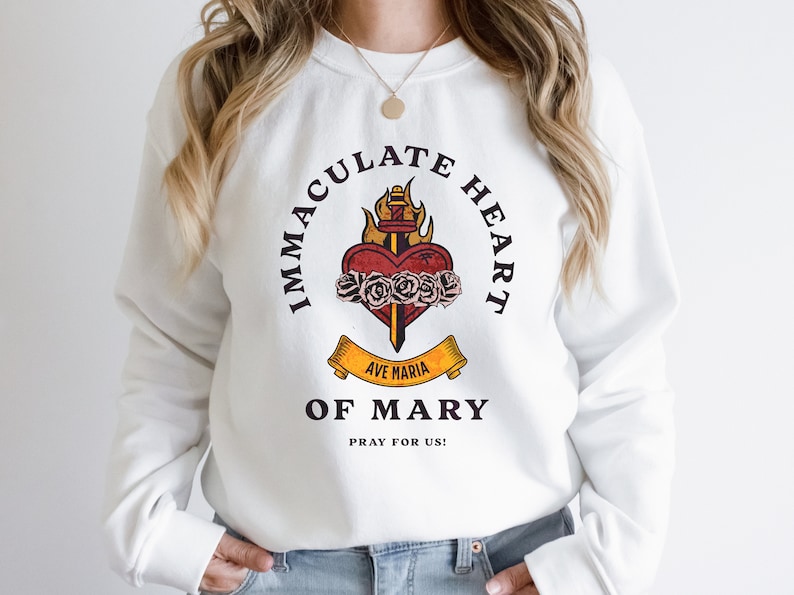 May include: White sweatshirt with a graphic print of a flaming heart with a sword and roses. The text around the heart reads "Immaculate Heart of Mary" and "Ave Maria" on a banner below the heart. The text below the graphic reads "Pray for Us!"