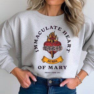 May include: Gray sweatshirt with a graphic print of a heart with a sword and flames. The text reads "Immaculate Heart of Mary" and "Ave Maria" with a banner below that says "Pray for Us!"