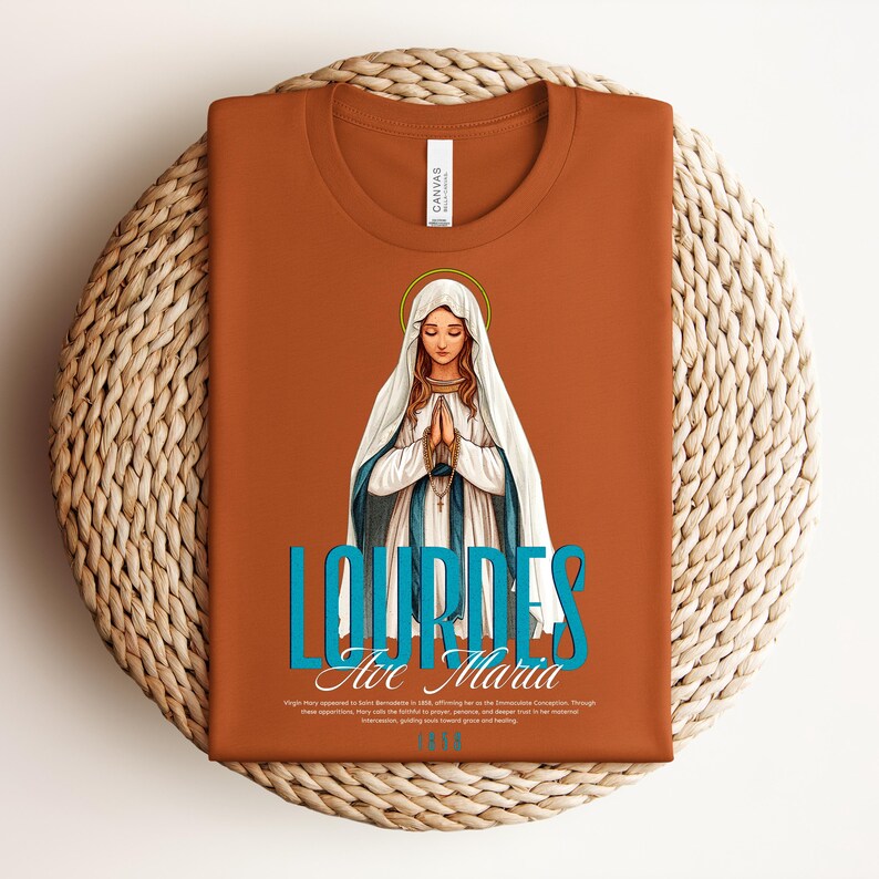 May include: A brown t-shirt with a blue and white graphic of the Virgin Mary in Lourdes, France. The text "Lourdes Ave Maria 1858" is printed below the image.