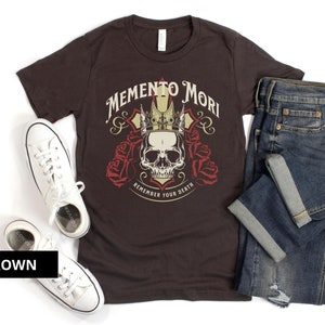 Pode incluir: Uma camiseta marrom com uma caveira usando uma coroa e o texto "Memento Mori Remember Your Death".