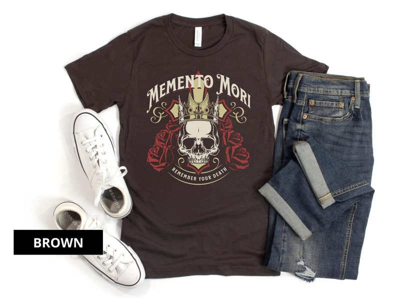 Pode incluir: Uma camiseta marrom com uma caveira usando uma coroa e o texto "Memento Mori Remember Your Death".