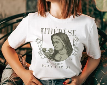 Puede incluir: Camiseta blanca con un estampado gráfico de Santa Teresa de Lisieux, una santa católica. El gráfico presenta un retrato de Teresa con hábito de monja, rodeado de rosas y el texto "SAINT THÉRÈSE OF LISIEUX PRAY FOR US".