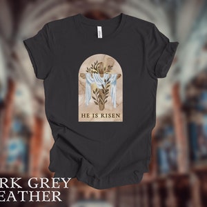 May include: Dark grey heather t-shirt with a graphic of a wooden cross with a white cloth draped over it. The graphic is set against a background of a marble archway with green leaves. The text "HE IS RISEN" is printed below the graphic.