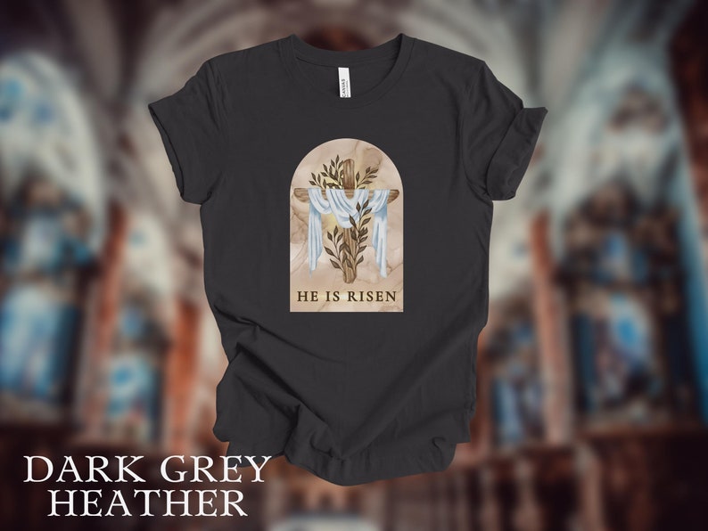 May include: Dark grey heather t-shirt with a graphic of a wooden cross with a white cloth draped over it. The graphic is set against a background of a marble archway with green leaves. The text "HE IS RISEN" is printed below the graphic.