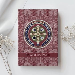May include: A book with a burgundy cover featuring a repeating pattern of religious symbols and text. The center has a circular emblem with a cross and Latin text. The name "FRANCIS PAUL" is printed at the bottom.