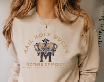 May include: A beige sweatshirt with the text "Hail Holy Queen" and a crown with a blue "M" in the center. The text "Mother of Mercy" is below the crown with the text "Our life, our sweetness and our hope" below that.
