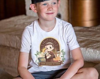 May include: White youth t-shirt with a cartoon illustration of Saint Anthony of Padua holding a baby Jesus. The illustration is surrounded by flowers and the text "Pray for us" and "Saint Anthony of Padua".