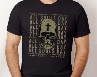 Puede incluir: Camiseta negra con un gráfico de calavera y el texto "All Souls Day" repetido varias veces. La calavera tiene una cruz en la frente. El texto "Remember, it is in dying that we are born to eternal life. Saint Francis of Assisi" está impreso debajo del gráfico.