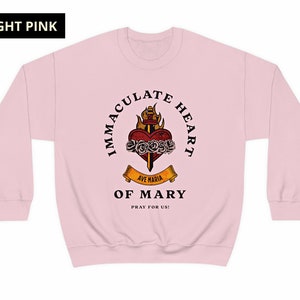 May include: A light pink sweatshirt with a graphic of the Immaculate Heart of Mary. The graphic features a heart with a sword, roses, and flames. The text "Immaculate Heart of Mary" is written in a circle around the graphic. The text "Ave Maria" is written on a banner below the graphic. The text "Pray for us!" is written below the banner.