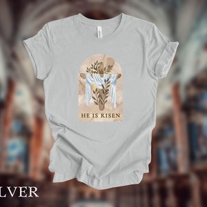 May include: A light gray t-shirt with a watercolor illustration of a cross with a crown of thorns and the text "He is Risen".