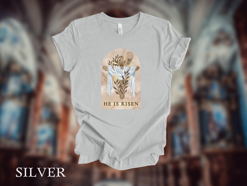 May include: A light gray t-shirt with a watercolor illustration of a cross with a crown of thorns and the text "He is Risen".