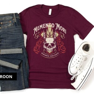 Pode incluir: Uma camiseta de cor vinho com um gr&aacute;fico de caveira e coroa e o texto "Memento Mori Remember Your Death".