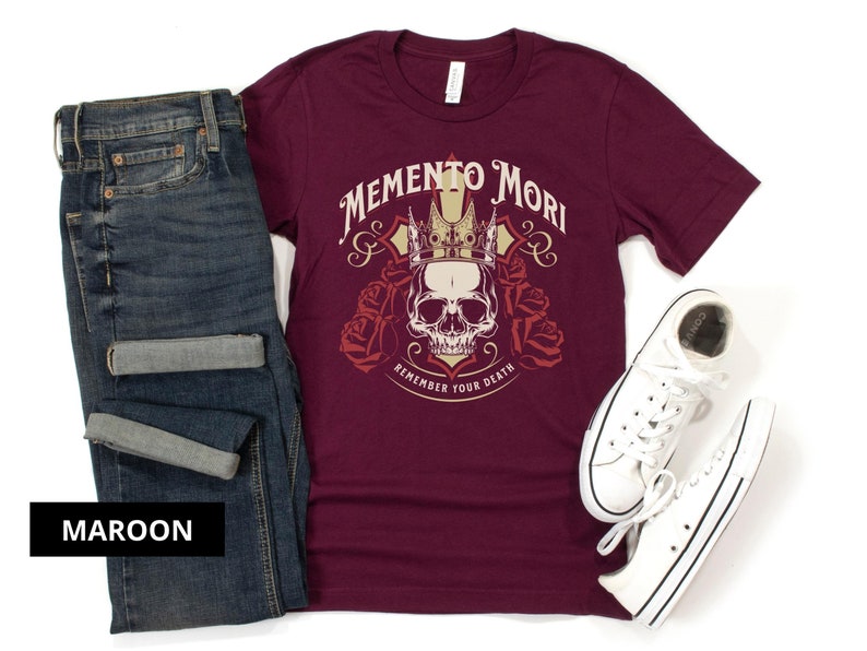 Pode incluir: Uma camiseta de cor vinho com um gr&aacute;fico de caveira e coroa e o texto "Memento Mori Remember Your Death".