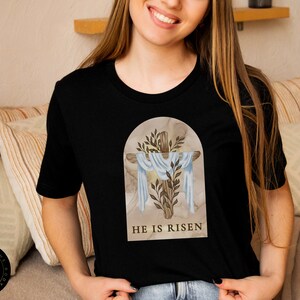 May include: Black t-shirt with a graphic of a cross with a white cloth draped over it. The cross is surrounded by green leaves and the text "HE IS RISEN" is printed below the cross.