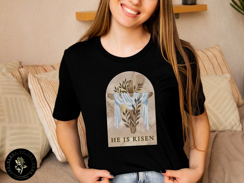 May include: Black t-shirt with a graphic of a cross with a white cloth draped over it. The cross is surrounded by green leaves and the text "HE IS RISEN" is printed below the cross.