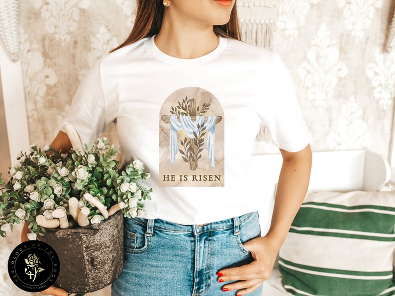 May include: White t-shirt with a graphic of a cross with a white cloth draped over it, surrounded by leaves and a marble background. The text "HE IS RISEN" is printed below the graphic.