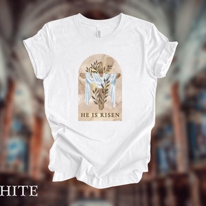 May include: White t-shirt with a graphic of a wooden cross with a white cloth draped over it. The cross is surrounded by green leaves and the text "He is Risen" is printed below the cross.