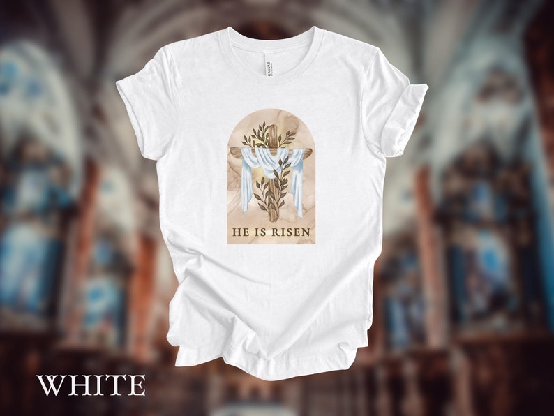 May include: White t-shirt with a graphic of a wooden cross with a white cloth draped over it. The cross is surrounded by green leaves and the text "He is Risen" is printed below the cross.