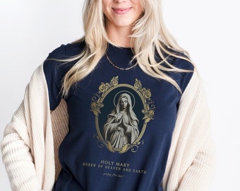 May include: A navy blue t-shirt with a gold illustration of the Virgin Mary in a frame. The text "Holy Mary Queen of Heaven and Earth pray for us!" is printed below the image.