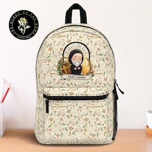 Peut inclure: Un sac à dos beige avec un motif répété de symboles religieux et le texte "Saint Charbel". Le sac à dos a une fermeture éclair noire, une poche avant et une poignée supérieure. L'élément principal est une illustration de Saint Charbel.