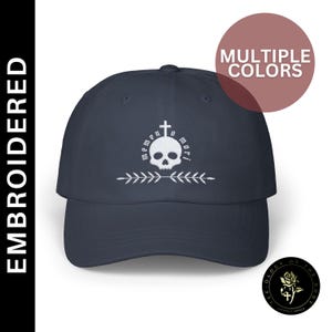 May include: A navy blue baseball cap with a white embroidered skull and crossbones design. The text "memento mori" is also embroidered in white on the cap.