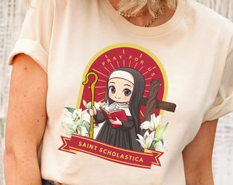 Op de afbeelding: Een beige T-shirt met een cartoon afbeelding van Sint Scholastica die een boek en een staf vasthoudt. De afbeelding is omringd door witte lelies en de tekst "Pray for Us" in een rode cirkel.