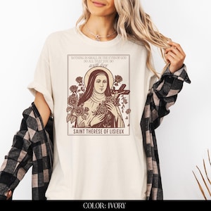 May include: Ivory-colored t-shirt featuring a sepia-toned graphic of Saint Th&eacute;r&egrave;se of Lisieux, surrounded by roses and holding a cross. The text on the shirt reads "NOTHING IS SMALL IN THE EYES OF GOD DO ALL THAT YOU DO with love".