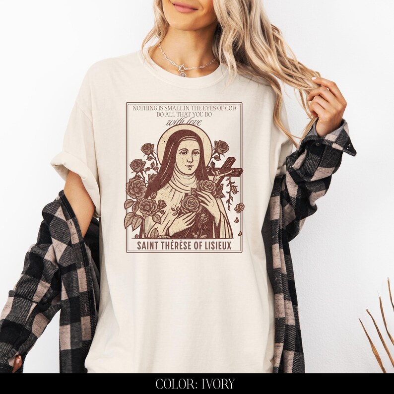 May include: Ivory-colored t-shirt featuring a sepia-toned graphic of Saint Th&eacute;r&egrave;se of Lisieux, surrounded by roses and holding a cross. The text on the shirt reads "NOTHING IS SMALL IN THE EYES OF GOD DO ALL THAT YOU DO with love".