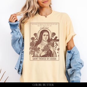 May include: Butter-colored t-shirt featuring a sepia-toned graphic of Saint Th&eacute;r&egrave;se of Lisieux, surrounded by roses and a cross. The text reads "NOTHING IS SMALL IN THE EYES OF GOD DO ALL THAT YOU DO with love."