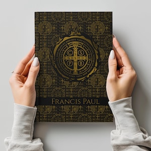 Puede incluir: Un libro con una cubierta negra y dorada que presenta un emblema circular detallado y patrones repetidos. El título del libro, "FRANCIS PAUL", se muestra en letras doradas. El libro está sostenido por dos manos.