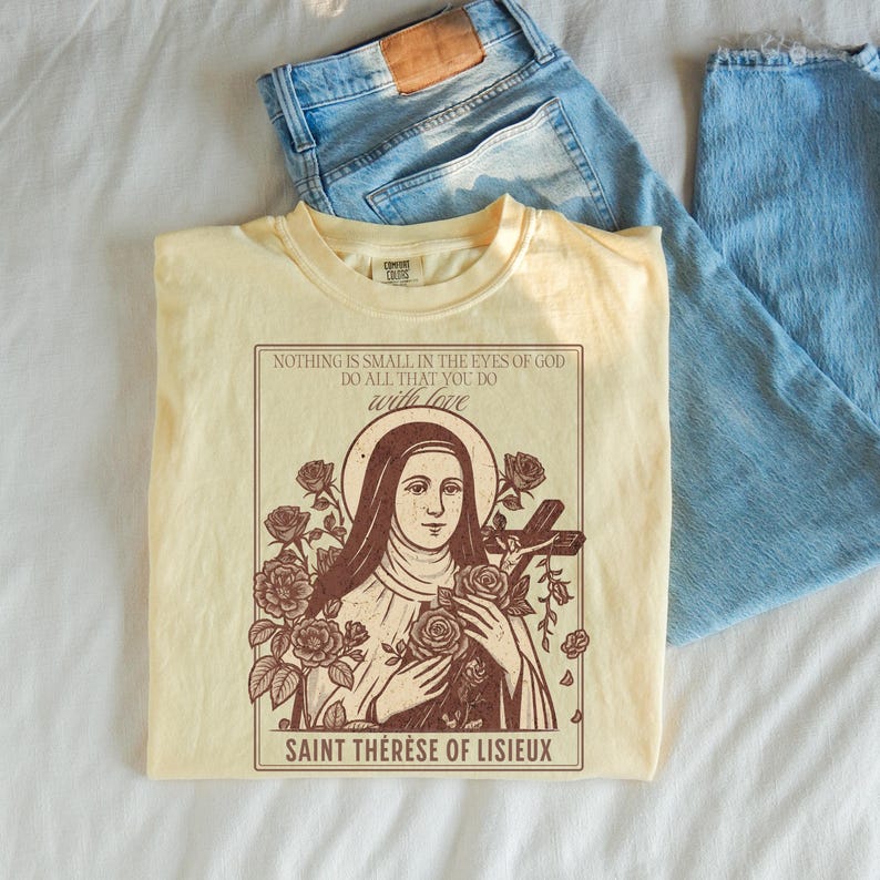 May include: Cream-colored t-shirt featuring a graphic of Saint Th&eacute;r&egrave;se of Lisieux, surrounded by roses and a cross. The shirt has text that reads "NOTHING IS SMALL IN THE EYES OF GOD DO ALL THAT YOU DO with love SAINT TH&Eacute;R&Egrave;SE OF LISIEUX." Light blue denim jeans are in the background.