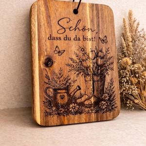 Könnte beinhalten: Holz-Schneidebrett mit dekorativem Blumendesign und dem deutschen Text "Schön, dass du da bist!" (Schön, dass du da bist!). Das Brett zeigt eingravierte Bilder von Blumen, Schmetterlingen, einer Gießkanne und Stiefeln.