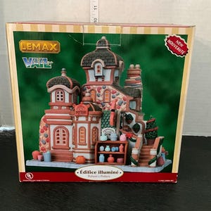 Puede incluir: Un edificio decorativo de cerámica iluminada de la colección Lemax Vail Village. El edificio presenta una tienda de cerámica con fachada roja y blanca, detalles de piedra y un techo detallado. La caja dice "Nuevo" y "Polson's Pottery."