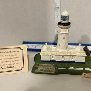 May include: A white lighthouse figurine on a green base with a blue ruler showing a height of approximately 6 inches. The base features text reading Macquarie N.S.W. Australia 542/9500 1997. Includes a certificate of authenticity and a small label.