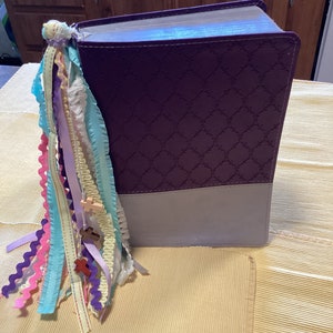 May include: A purple and grey hardback book with a decorative embossed pattern. The book has a colourful ribbon bookmark with small wooden crosses attached.