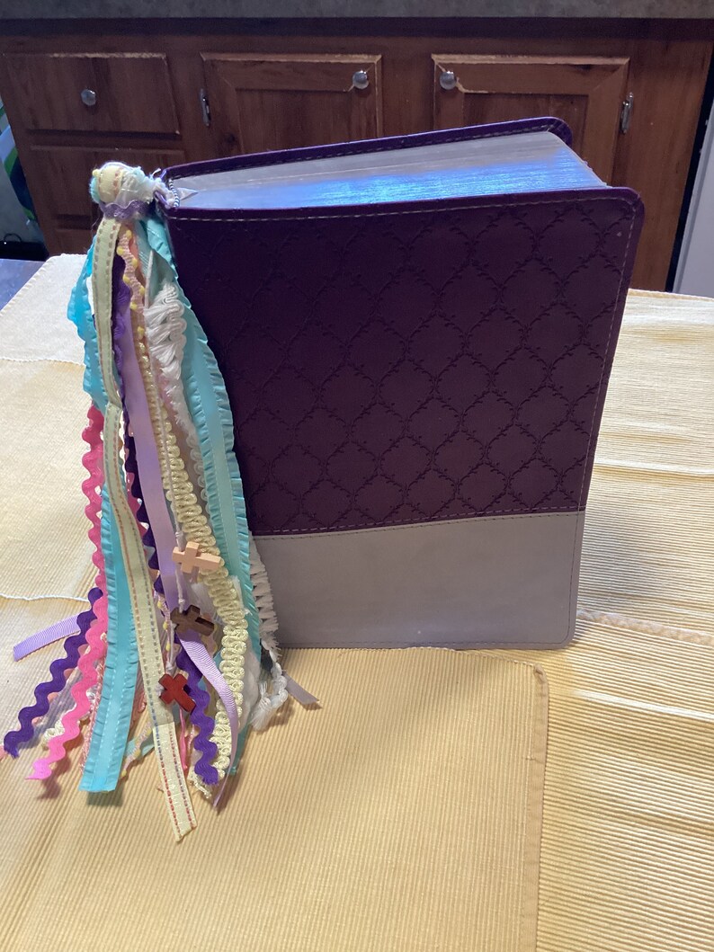 May include: A purple and grey hardback book with a decorative embossed pattern. The book has a colourful ribbon bookmark with small wooden crosses attached.