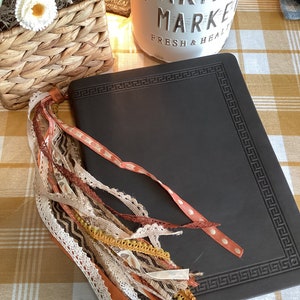 May include: A black leather journal with a decorative border and a tassel made of various fabrics in shades of brown, orange, and cream. The journal is lying on a checkered tablecloth.