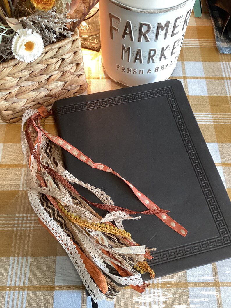 May include: A black leather journal with a decorative border and a tassel made of various fabrics in shades of brown, orange, and cream. The journal is lying on a checkered tablecloth.