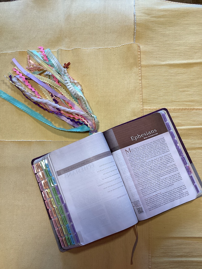 May include: An open Bible with a purple cover and colourful tabs. The Bible is open to the book of Ephesians. The text on the page reads "For by grace you have been saved through faith."