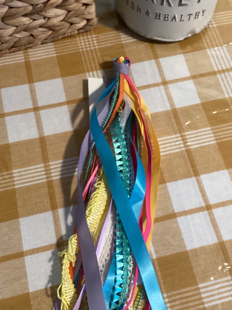 May include: A colourful assortment of ribbons and trims tied together in a bundle. The ribbons are in shades of blue, green, yellow, pink, and purple.