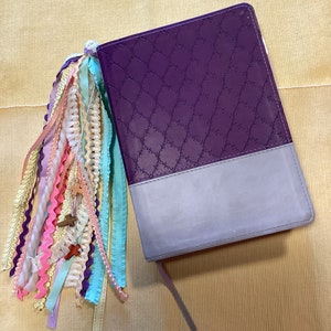 May include: A purple and grey journal with a decorative embossed pattern on the cover. The journal has a long tassel made of colourful ribbons attached to the spine.